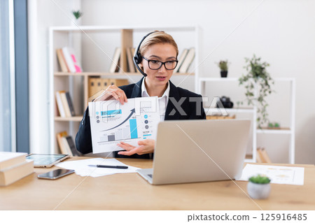 Professional businesswoman in her 30s showcasing chart during video conference call in office. Professional businesswoman in her 30s showcasing chart during video conference call in office. 125916485