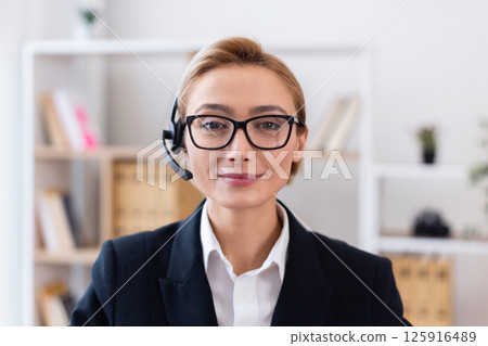 Smiling young caucasian woman wearing glasses and business suit with headset, standing in modern office setting providing customer service. Depicts professionalism, communication skills Smiling young caucasian woman wearing glasses and business suit with headset, standing in modern office setting providing customer service. Depicts professionalism, communication skills 125916489