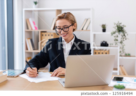 Middle-aged woman in professional outfit working in office writing notes and using laptop. Workspace is modern and organized with shelves and plants, creating a productive and positive atmosphere. 125916494