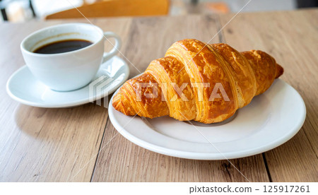 A delicious, buttery croissant is served on a white plate alongside a cup of black coffee, set on a wooden breakfast table, breakfast, coffee break, warm morning coffee 125917261
