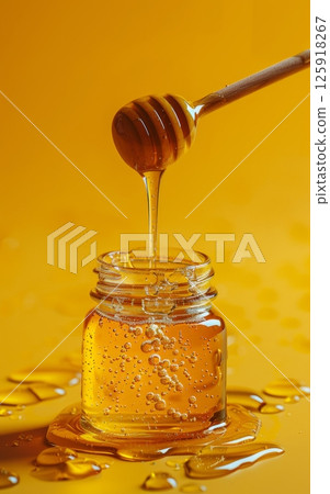 A Glass Jar with a Lid on a Solid Color Background with Honey and a Stirring Stick 125918267