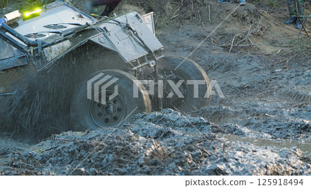 A close-up of a rugged, off-road vehicle splashing through a muddy course during a race. The vehicle has large tires and is partially obscured by mud and water 125918494