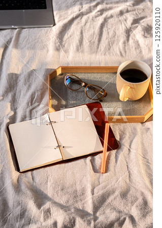 Open Notebook on Bed with Coffee Cup, Glasses and Laptop. Cozy Morning Scene for Writing and Planning. Open Notebook on Bed with Coffee Cup, Glasses and Laptop. Cozy Morning Scene for Writing and Planning. 125920110