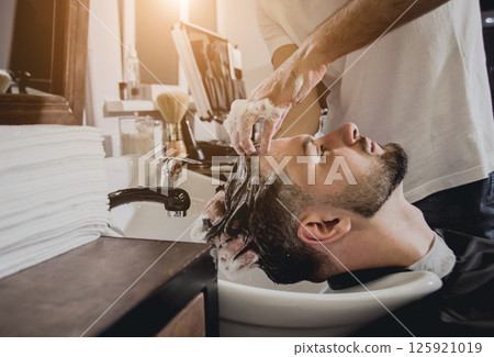 Young man with trendy haircut at barber shop. Barber washes customer head. 125921019