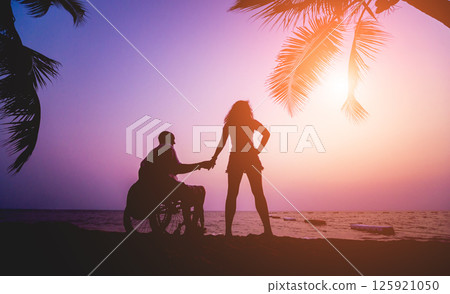 Disabled man in a wheelchair with his wife on the beach. Silhouettes at sunset 125921050