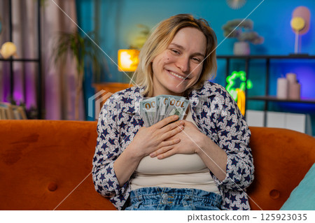 Happy excited girl winning online casino lottery game waving dollar bill money fan sitting on couch 125923035
