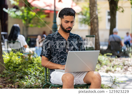 Indian man in headphones working online distant job with laptop browsing website chatting outdoors Indian man in headphones working online distant job with laptop browsing website chatting outdoors 125923229