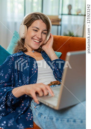 Happy woman in wireless headphones lying on home couch listening music working on laptop relaxing 125923242