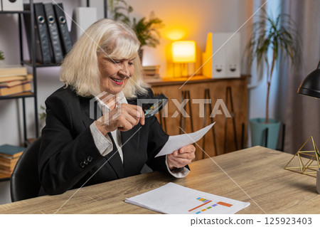 Senior businesswoman looks through magnifying glass, examine reading documents at home office desk 125923403