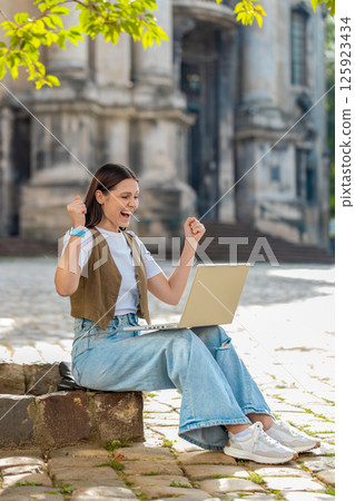 Overjoyed happy young woman working on laptop screaming in delight raises hands in city street Overjoyed happy young woman working on laptop screaming in delight raises hands in city street 125923434