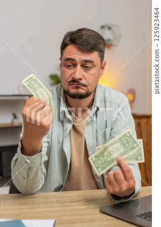 Upset Caucasian man freelancer counting dollar cash insufficient amount of money sitting at table 125923464
