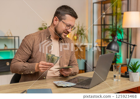 Rich happy Caucasian man counting money cash using laptop calculate domestic bills at home table 125923468