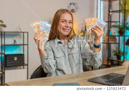 Excited businesswoman freelancer holding fan of euro cash and dancing sitting at home office table Excited businesswoman freelancer holding fan of euro cash and dancing sitting at home office table 125923472