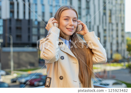 Caucasian mature woman taking off wireless headphones looking at camera and smiling on city street 125923482