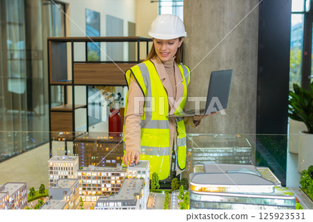 Confident architect in hardhat using laptop while analyzing office complex model at workplace 125923531