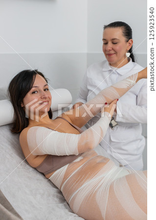 Cosmetologist warping woman hand with compression elastic bandage body mask regeneration weight loss Cosmetologist warping woman hand with compression elastic bandage body mask regeneration weight loss 125923540