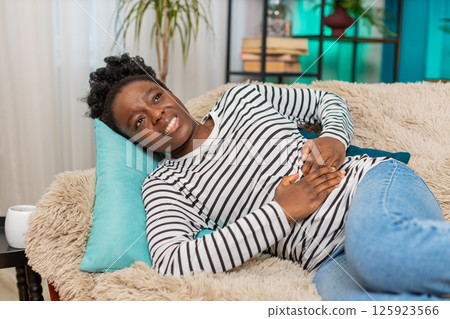 African American woman feeling stomach pain, holding abdomen, lying exhausted on couch indoors African American woman feeling stomach pain, holding abdomen, lying exhausted on couch indoors 125923566