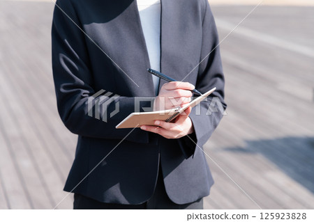 A young woman in a suit sitting on a bench outdoors and taking notes 125923828
