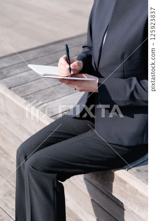 A young woman in a suit sitting on a bench outdoors and taking notes 125923837