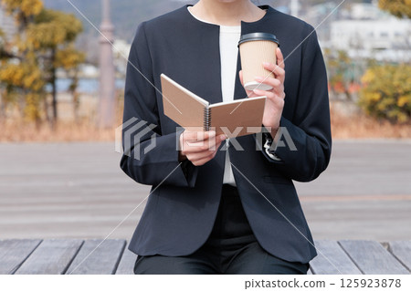 A young woman in a suit drinking coffee and looking at a notepad 125923878