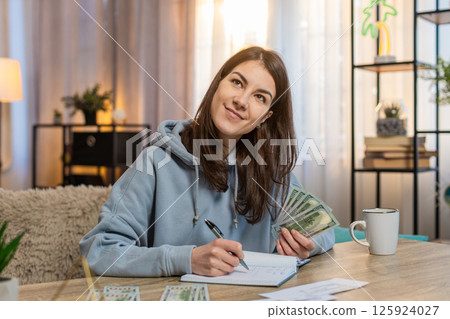 Young woman sitting at table looking at receipts and counting money while making shopping list 125924027