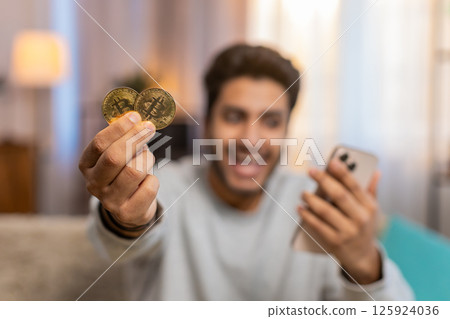 Young Indian man celebrating cryptocurrency win, showing bitcoin after checking exchange charts 125924036