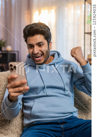 Excited Indian man holding smartphone celebrating online game win sitting on sofa in living room 125924038