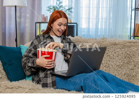 Woman sitting on couch watching movie on laptop eating popcorn laughing emotionally with excitement 125924095