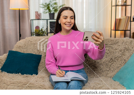 Young woman on sofa wearing headphones watching video lesson on mobile phone taking study notes Young woman on sofa wearing headphones watching video lesson on mobile phone taking study notes 125924105
