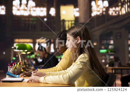 Two young students preparing for a lecture and writing ideas in notebooks at a study desk in a library, creating a reflective essay as homework. Girls focused on their school work. 125924124