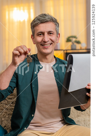 Young man smiles proudly holding keys documents after signing contract for new apartment at home 125924139