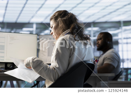 Woman overseeing server room, reading documentation paperwork, troubleshooting hardware. Supervisor doing inspection in server hub, reading technical files at computer desk 125924146