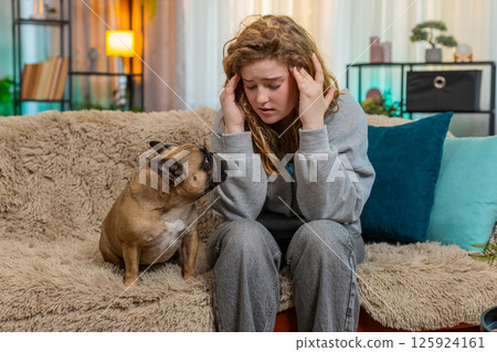 Caucasian woman with headache sits on home sofa while pug dog cuddles face and shows empathy gently 125924161