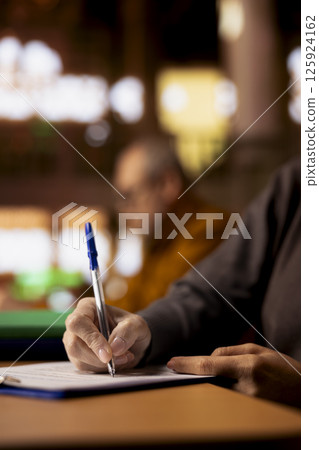 Mature academic person prepares documents for her university coursework, transcribing from literature and textbooks. Scholar focused on research and education in a public library. Close up. Mature academic person prepares documents for her university coursework, transcribing from literature and textbooks. Scholar focused on research and education in a public library. Close up. 125924162