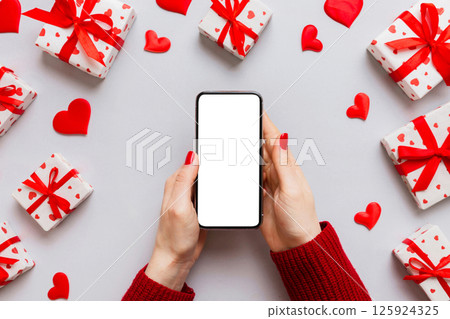 Woman hand holding mobile phone with blank screen on colored background with hearts, valentine day concept top view flat lay 125924325
