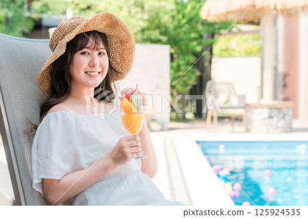 年輕的亞洲女性坐在泳池邊的躺椅上喝熱帶果汁 125924535