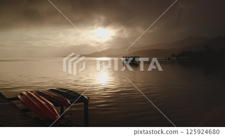 Dramatic sunrise over tranquil lagoon with colorful kayaks resting on wooden dock and boat moored near pier, creating a serene and picturesque morning scene. Tropical landscape on exotic island beach 125924680