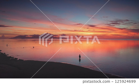 Breathtaking aerial view of a vibrant sunset reflected in calm ocean waters, with a lone man standing on the shoreline, creating a serene and picturesque scene in a tropical paradise 125924699