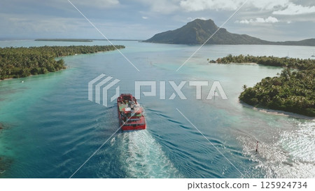 Aerial drone of red cargo ship sailing through crystal clear waters of Maupiti lagoon, tropical islands and mount Teurafaatui in background. Delivery bulk carrier to exotic remote places 125924734