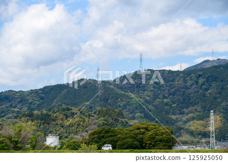 A scenic landscape featuring lush green mountains under a partly cloudy sky. Tall electricity transmission towers line the ridges A scenic landscape featuring lush green mountains under a partly cloudy sky. Tall electricity transmission towers line the ridges 125925050