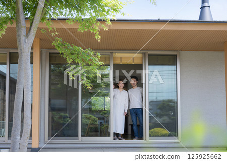 A couple looking at the garden from the veranda or under the veranda of a house surrounded by greenery, looking at the camera A couple looking at the garden from the veranda or under the veranda of a house surrounded by greenery, looking at the camera 125925662