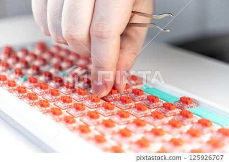 Hand installing red hot-swap switch into mechanical keyboard plate with tweezers during assembly Hand installing red hot-swap switch into mechanical keyboard plate with tweezers during assembly 125926707