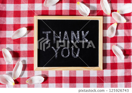 Small blackboard with Thank you message conveying gratitude on red checkered tablecloth and petals 125926743