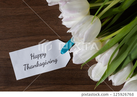 Happy Thanksgiving greeting tag placed near white tulips on a wooden surface, perfect for holiday celebration messages 125926768