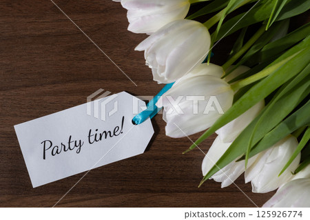 Tag with Party time text attached to white tulips on wooden background ideal for party invitations event announcements celebration messages or festive spring promotions 125926774