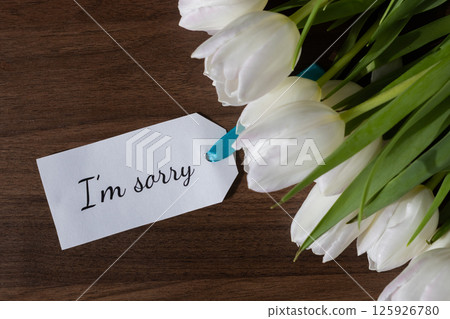 White tulip bouquet with an I'm sorry message note conveying apology and sympathy on dark wood, suitable for difficult emotional communication needs 125926780