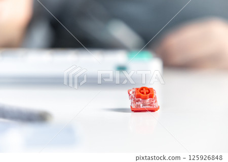 Single mechanical keyboard switch placed on white desk surface in front of blurred keyboard Single mechanical keyboard switch placed on white desk surface in front of blurred keyboard 125926848