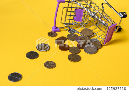 Mini shopping cart overturned with scattered euro coins on vibrant yellow background symbolizing loss Mini shopping cart overturned with scattered euro coins on vibrant yellow background symbolizing loss 125927538