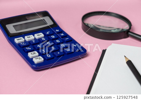Blue calculator, magnifying glass, and notebook with pencil on pink background symbolizing auditing 125927548