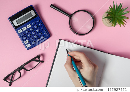Hand writing in notebook with calculator, magnifying glass, glasses and plant on pink workspace 125927551